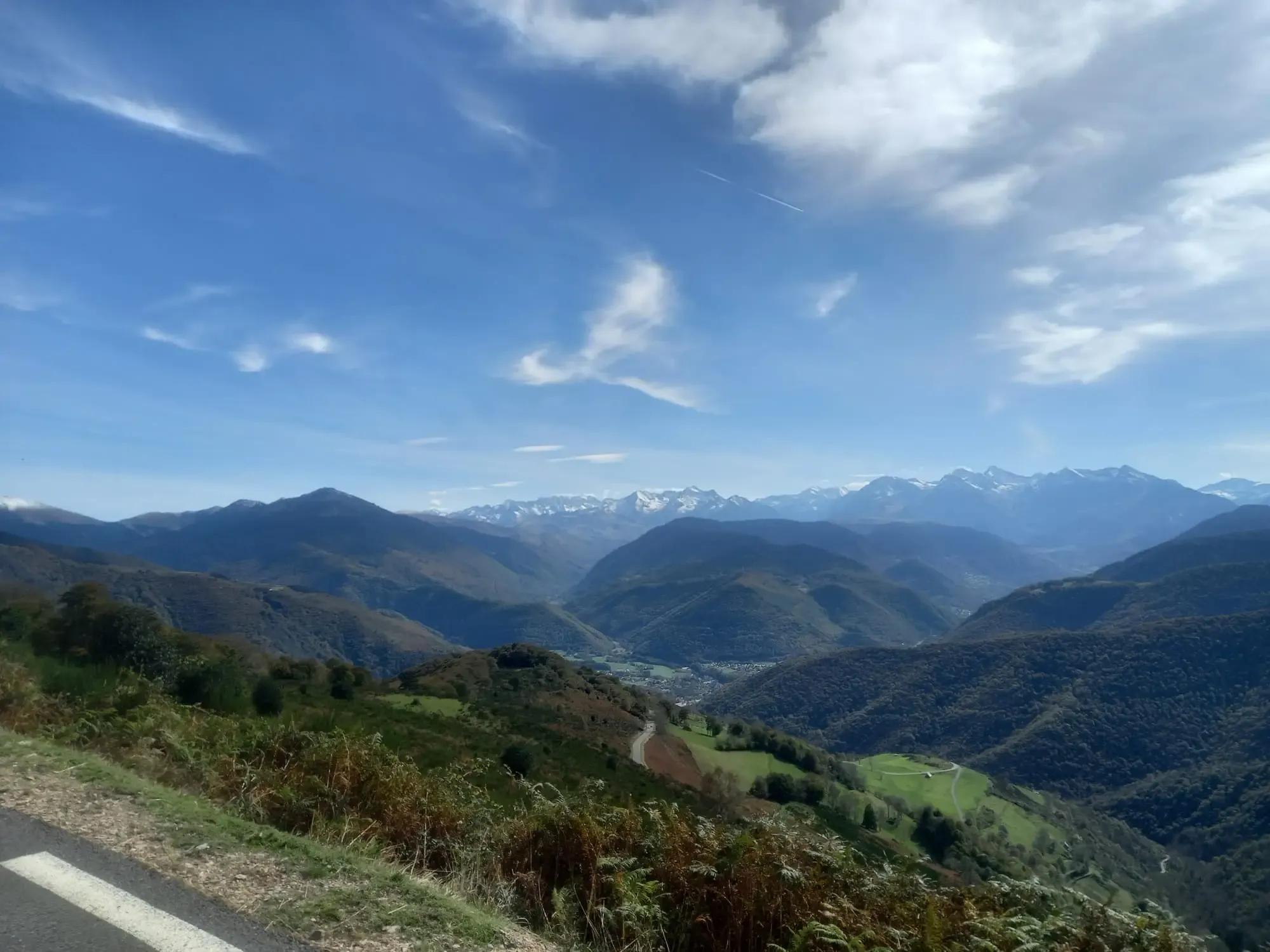 Vaste panorama des Pyrénées de l'Arbizon et du massif de Neouvielle depuis la route d'ascension du Col d'Aspin (côté Campan). La route départementale sinueuse est visible dans la vallée boisée au premier plan, avec des montagnes enneigées et un ciel bleu clair parsemé de nuages légers en arrière-plan.